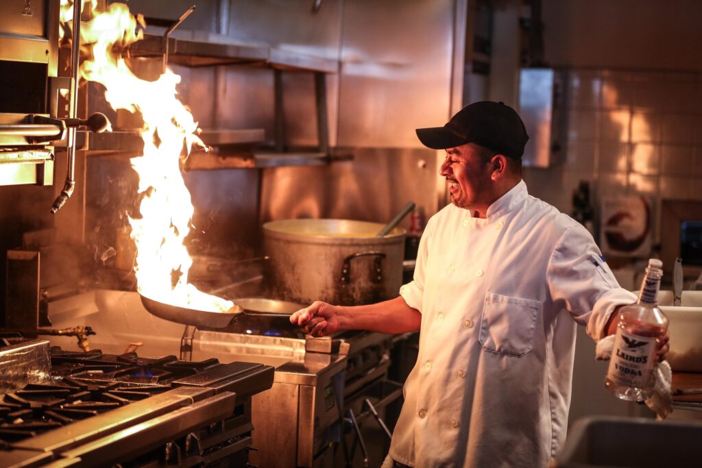 A chef with a white jacket and black hat holding a vodka bottle. Serving up food from a pan with a flame. Algorithms serve up content like a chef serves up food.